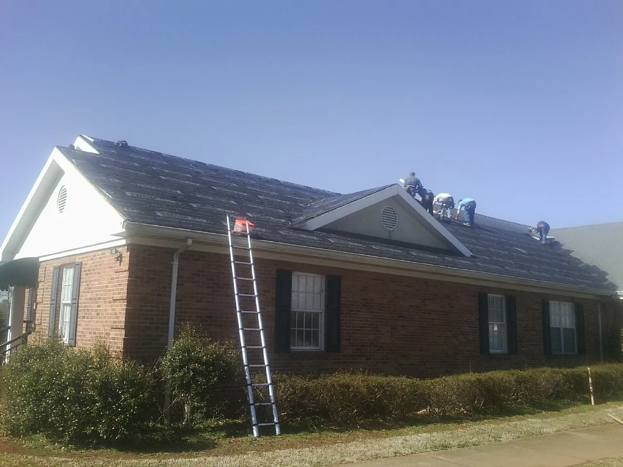 Crew tearing shingles off a brick ranch roof with a ladder set — Metal Roof Repair work by our crew in Olustee