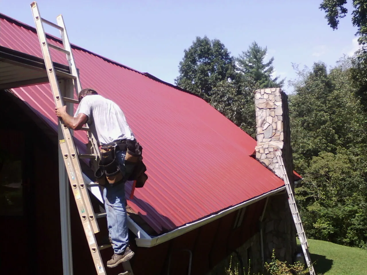 Roofer on a ladder beside a red metal roof and stone chimney — Soffit & Fascia Repair by Roofing Brothers in Olustee